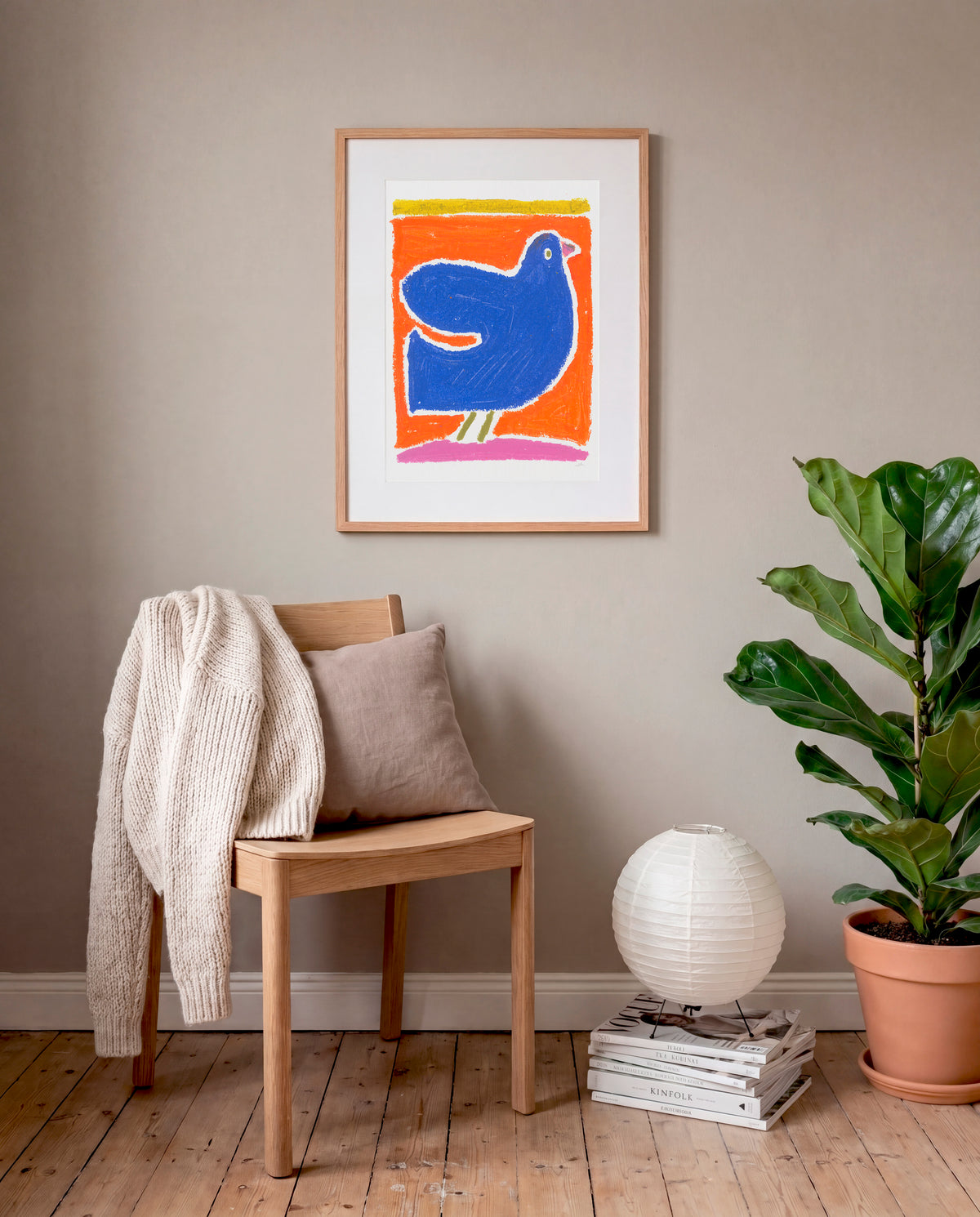 Het werk van Ella "Vogel" op de muur in een eiken frame met stoel en plant.